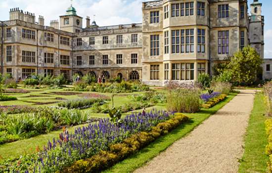 A view of Audley End House underneath a blue sky overlooking the gardens with rows of colourful flowers.