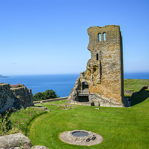 A ruined castle tower surrounded by grass with the sea in the distance on a sunny day (Scarborough Castle)