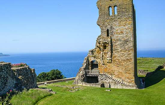 A ruined castle tower surrounded by grass with the sea in the distance on a sunny day (Scarborough Castle)