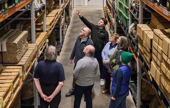 A man stands in front of a group of people in a storeroom pointing at one of the shelves