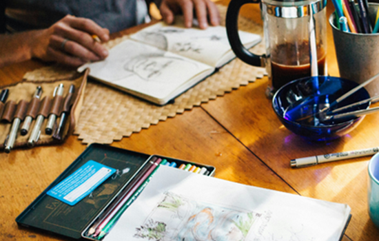A person seated at a wooden table sketching in an open notebook, surrounded by art supplies including pens, coloured pencils, a roll-up pen case, a bowl with brushes, and a French press of coffee.