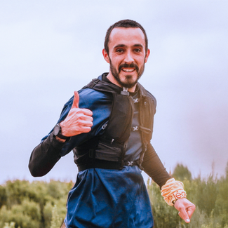 A person running outdoors on a trail, giving a thumbs‑up gesture, wearing a blue long‑sleeve top and a running vest, with greenery and an overcast sky in the background.
