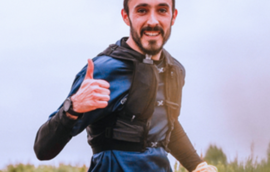 A person running outdoors on a trail, giving a thumbs‑up gesture, wearing a blue long‑sleeve top and a running vest, with greenery and an overcast sky in the background.